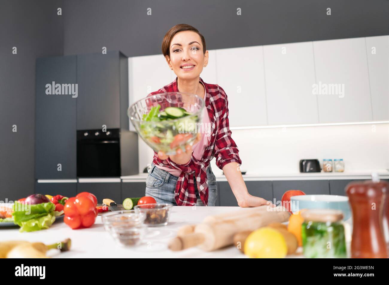 Glückliche junge Hausfrau hält eine Salatbäckerei und zeigt sie vor der Kamera, die für ein Familienessen vorbereitet ist, oder die Mädchennacht, die im Kit steht Stockfoto