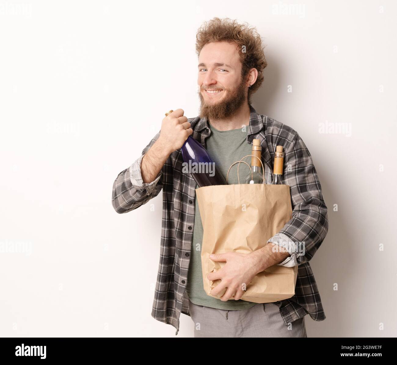 Zurück vom Einkaufen mit einem Papierbeutel mit Flaschen Wein lächelnder junger Mann. Hübscher junger Mann mit lockigen Haaren in olivfarbenem T-shi Stockfoto