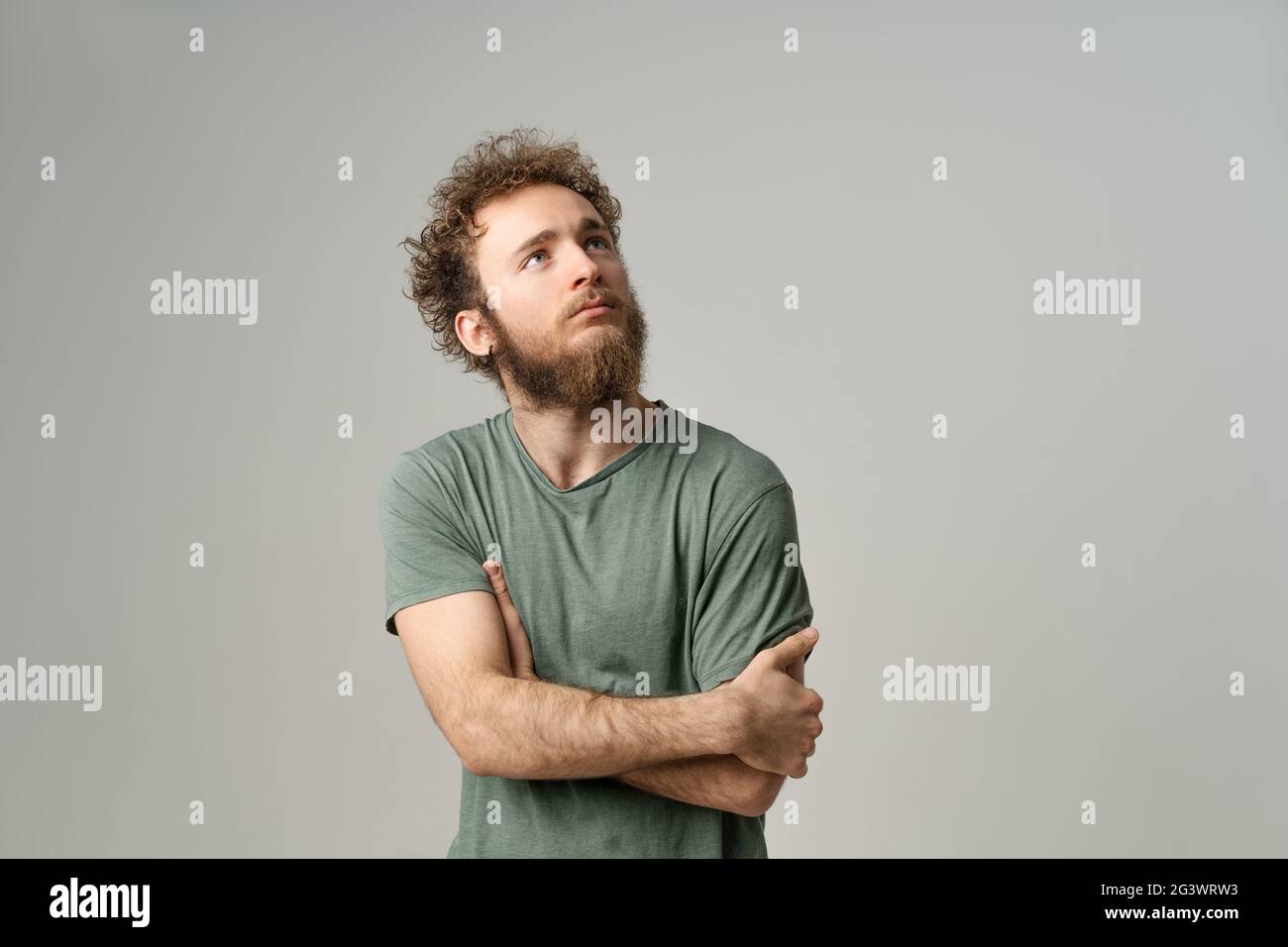 Porträt von nachdenklichen tief in Gedanken jungen Mann mit gefalteten Händen. Attraktiver Mann mit lockigem Haar und Bart in olivfarbener T-Shirt-Optik Stockfoto