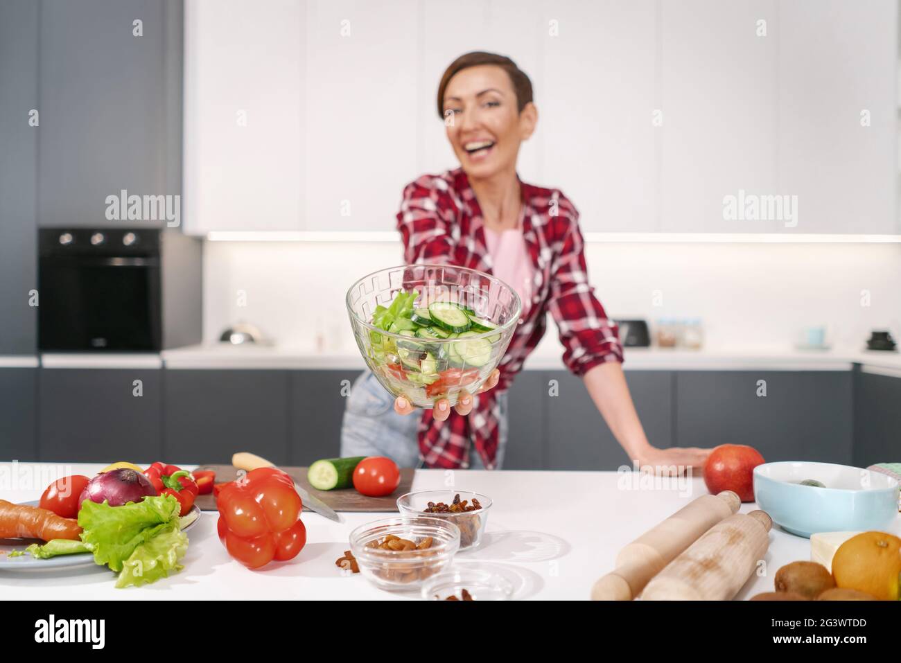 Glückliche junge Hausfrau hält eine Salatbäckerei und zeigt sie vor der Kamera, die für ein Familienessen vorbereitet ist, oder die Mädchennacht, die im Kit steht Stockfoto