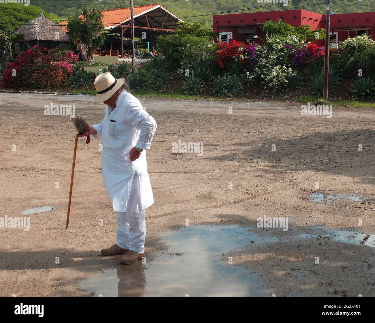 Ein Tourist in weißer Kleidung (Kurta), einem Stock und einem Strohhut, der vor einer Reihe von Häusern steht. Er prüft seine Tasche. Karibischer Urlaub. Stockfoto