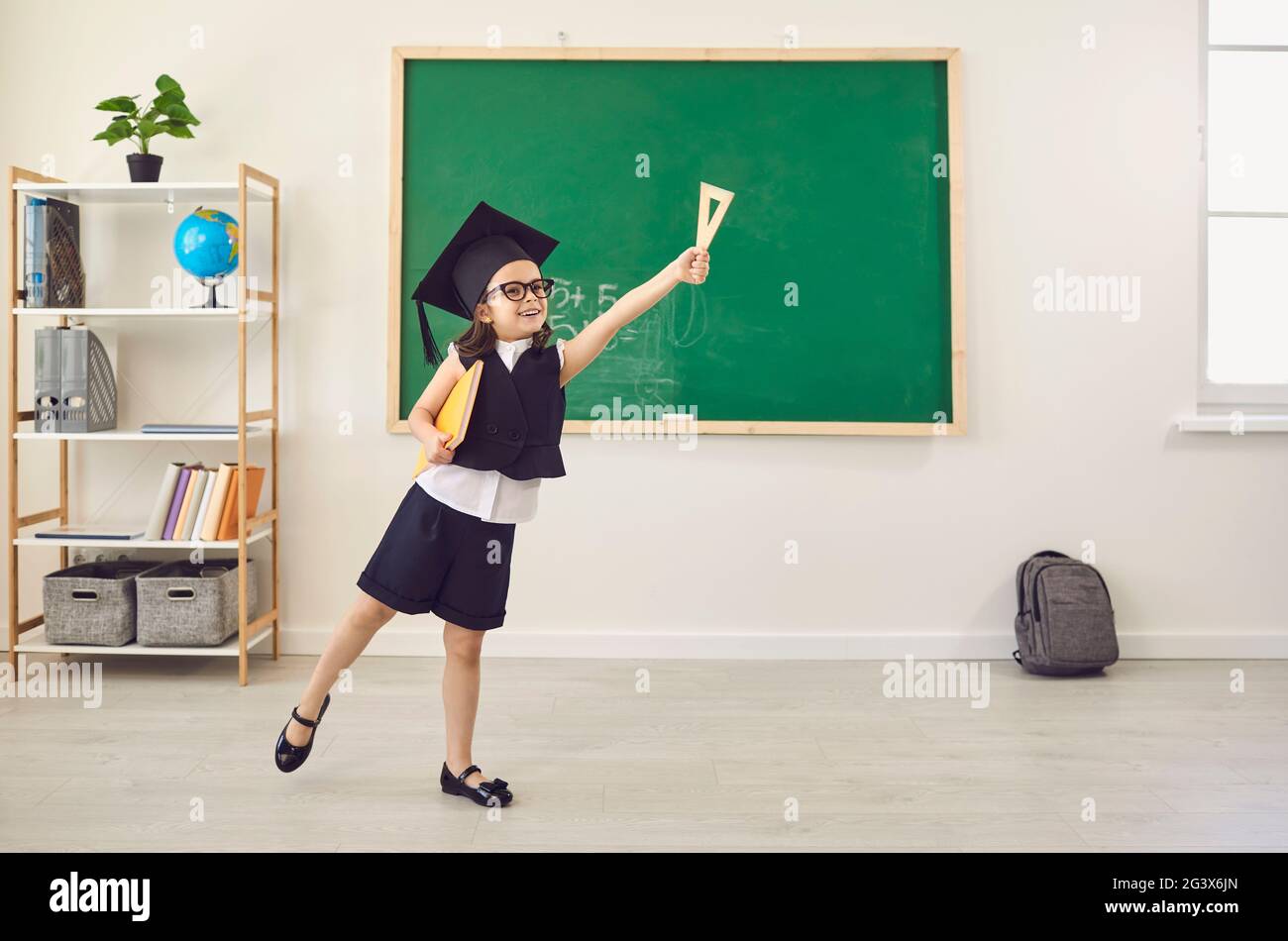 Zurück zur Schule. Das Schulmädchen in einer Abschlussmütze hob fröhlich ihre Hand im Lassroom nach oben. Stockfoto