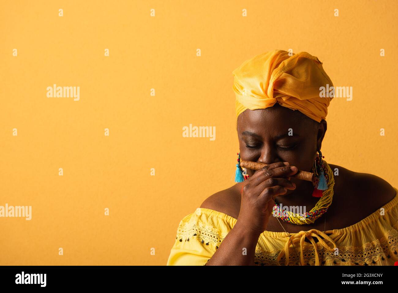 afroamerikanische Frau mittleren Alters in gelbem Turban riecht nach Zigarre mit geschlossenen Augen auf gelb Stockfoto