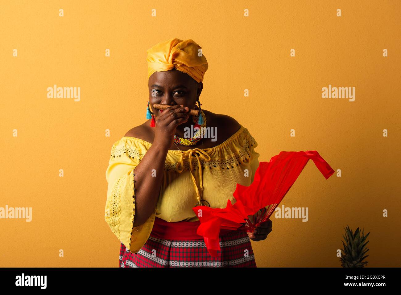 afroamerikanische Frau mittleren Alters in gelbem Turban und hellen Kleidern, die auf Gelb nach Zigarren riechen Stockfoto