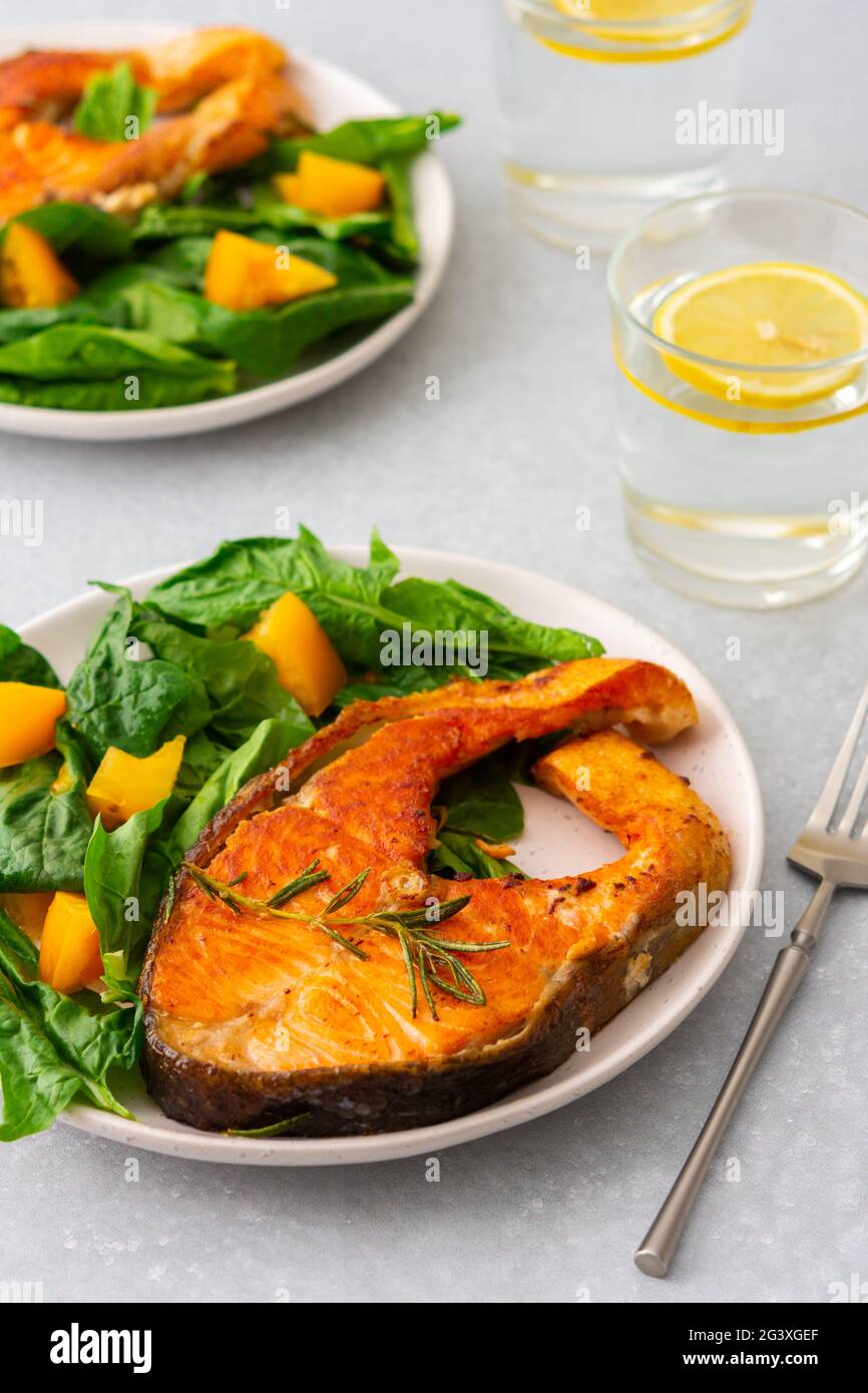 Gebratenes Lachssteak mit Spinat und gelben Tomaten auf einem Teller auf grauem Grund, ein Glas Wasser mit Zitrone, ein herzhaftes Mittagessen mit gesundem rotem Fisch Stockfoto
