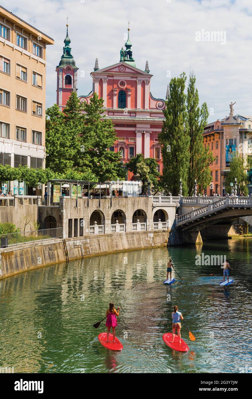 Ljubljana, Slowenien. Eine Gruppe junger Leute paddeln auf Paddel-Surfbrettern den Fluss Ljubljanica hinunter. Barocke Franziskanerkirche der Verkündigung Stockfoto