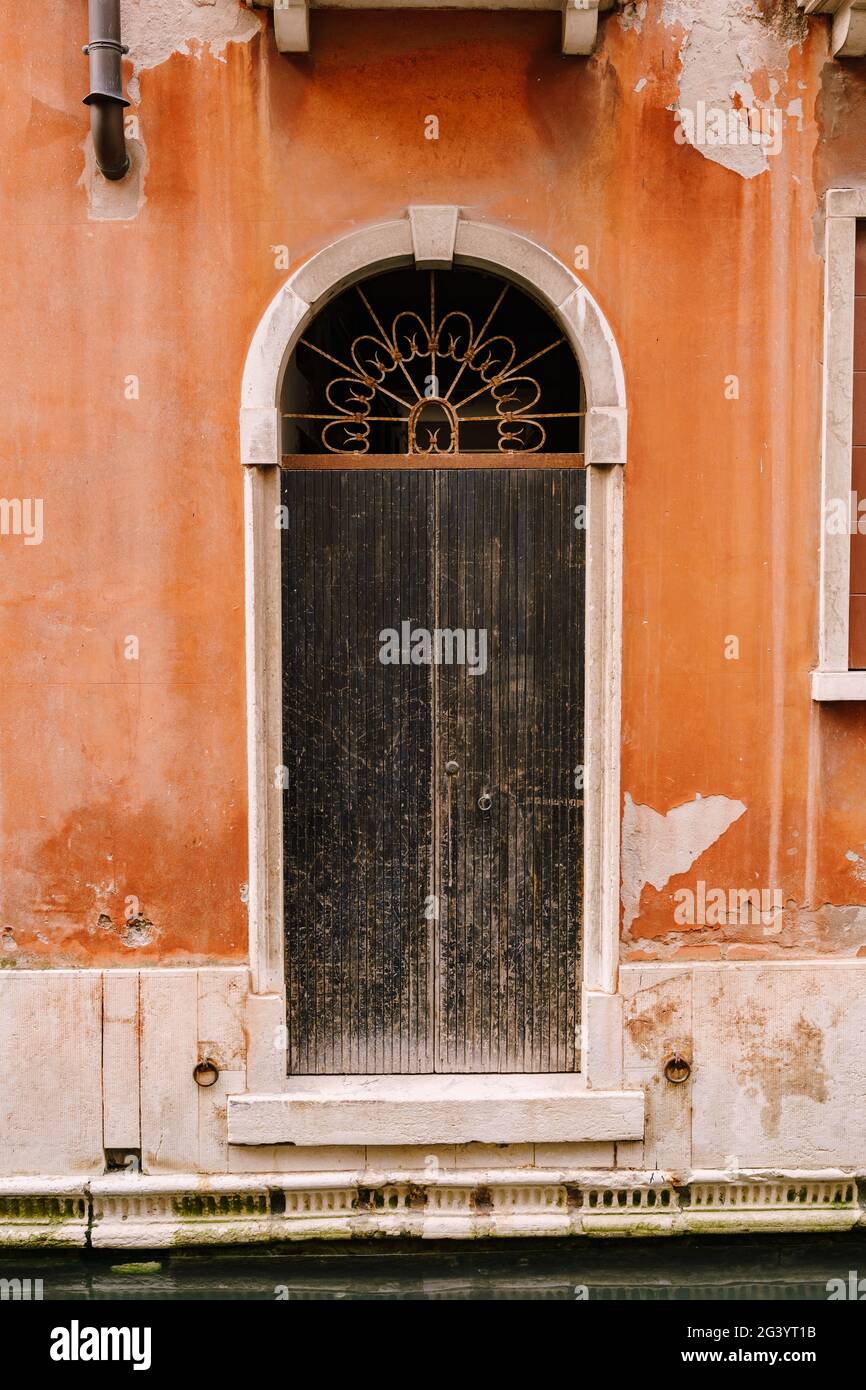 Nahaufnahmen von Gebäudefassaden in Venedig, Italien. Eine alte schwarze, zerkratzte Tür, in einer gewölbten Tür, mit einem rostig geschmiedeten Kühlergrill A Stockfoto
