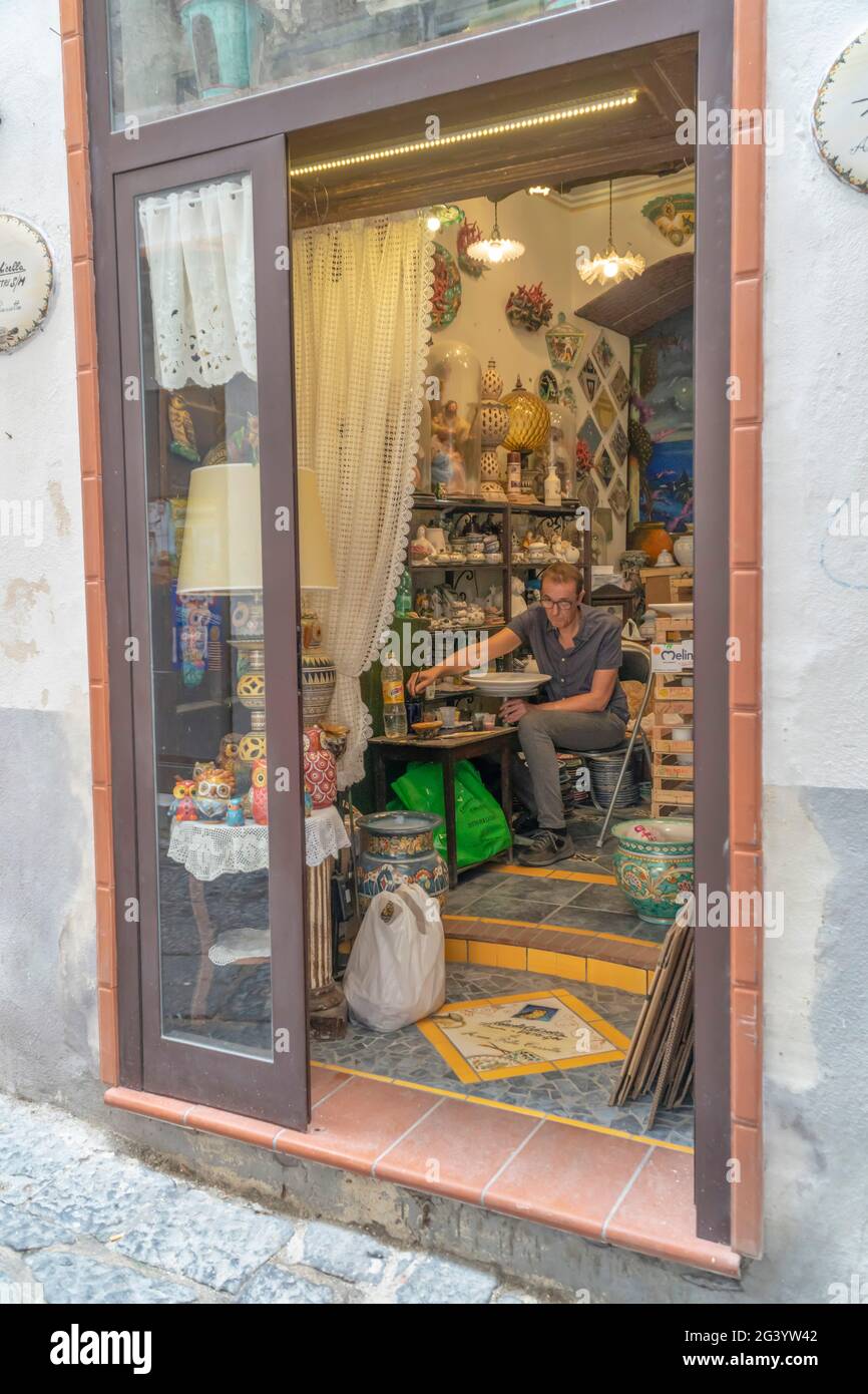 Eine Kunsthandwerkerwerkstatt in Vietri - Italien. Ein Handwerker malt einen Tonteller. Handgemachte Souvenirs. Viele Töpferobjekte an den Schrankregalen. Stockfoto