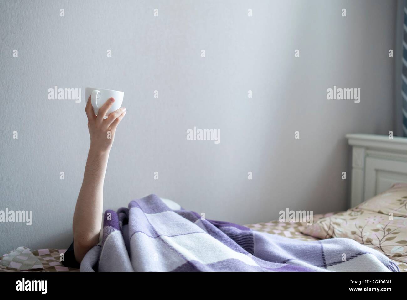 Erhobene Hand einer Person im Bett, die am frühen Morgen eine Tasse Tee hielt Stockfoto