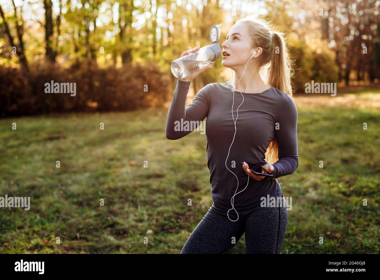 Fitness im Park, Mädchen trinken Wasser, halten Smartphone und Kopfhörer in der Hand. Stockfoto