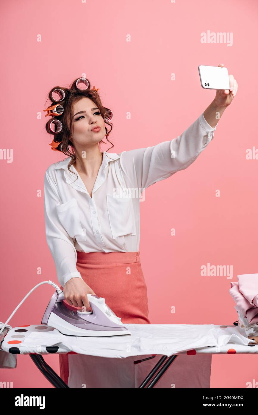 Vor einem rosa Hintergrund arbeitet eine wunderschöne Hausfrau mit Lockenstab Kleidung auf dem Bügelbrett und macht ein Selfie am Telefon. Nettes Mädchen doin Stockfoto