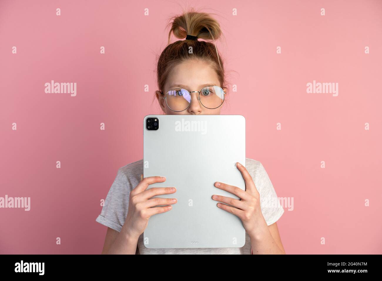 Attraktives, blondes Mädchen mit Tablet und Brille auf rosa Hintergrund. Der süße Teenager verwendet eine Tablette, sie bedeckte ihr Gesicht mit einer Tablette, nur ihre Augen Stockfoto