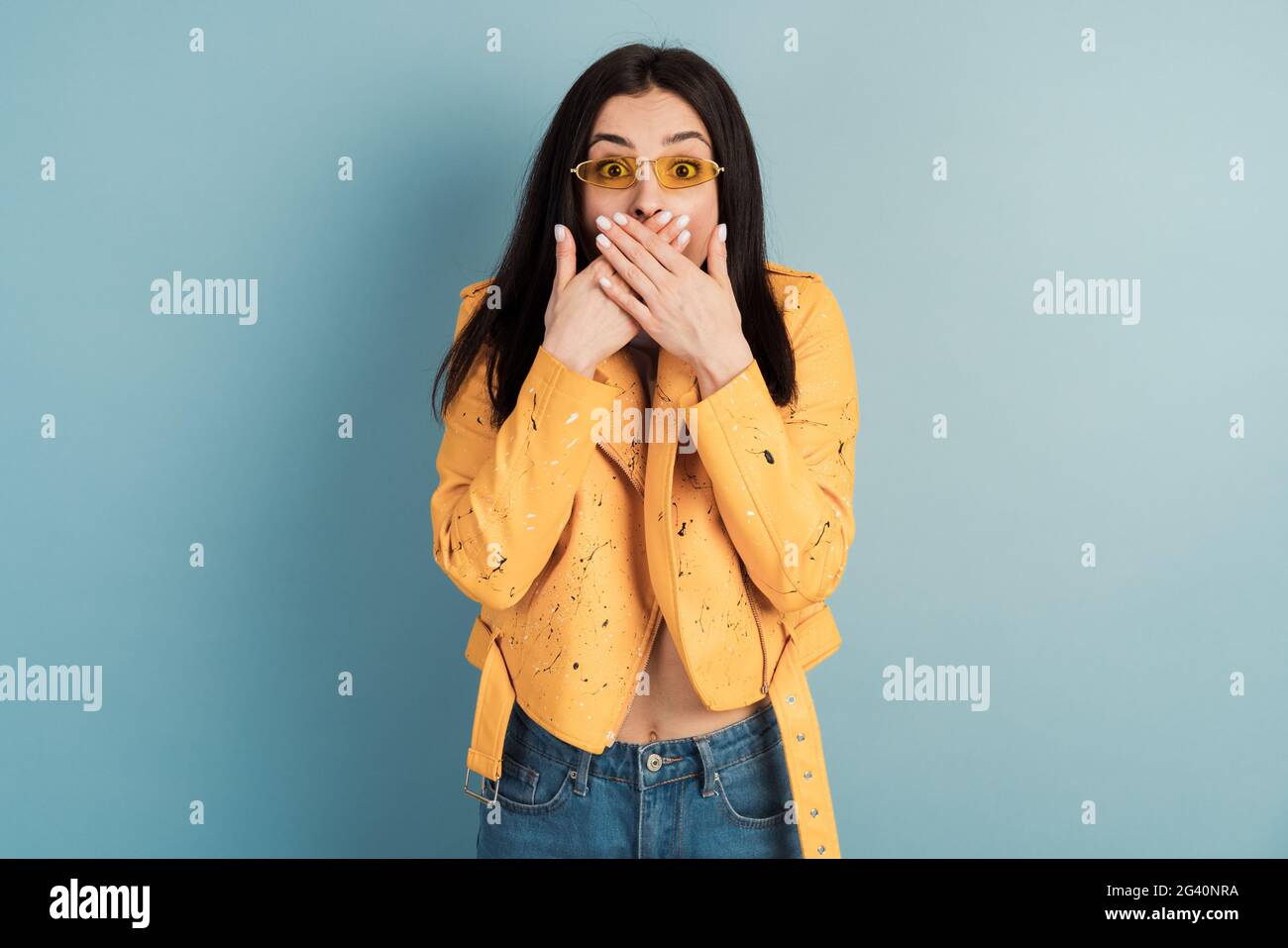 Eine junge Frau in orangefarbener Jacke auf blauem Hintergrund bedeckt ihren Mund mit beiden Händen. Das Konzept des Geheimnisses. Stockfoto