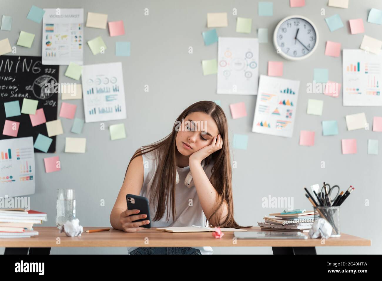 Müde, gelangweilte Mädchen mit einem Telefon an einem Tisch auf einem Hintergrund von grauen Wand mit Aufklebern sitzen. Lernkonzept., müde, gelangweilte Mädchen mit einem Telefon sittin Stockfoto