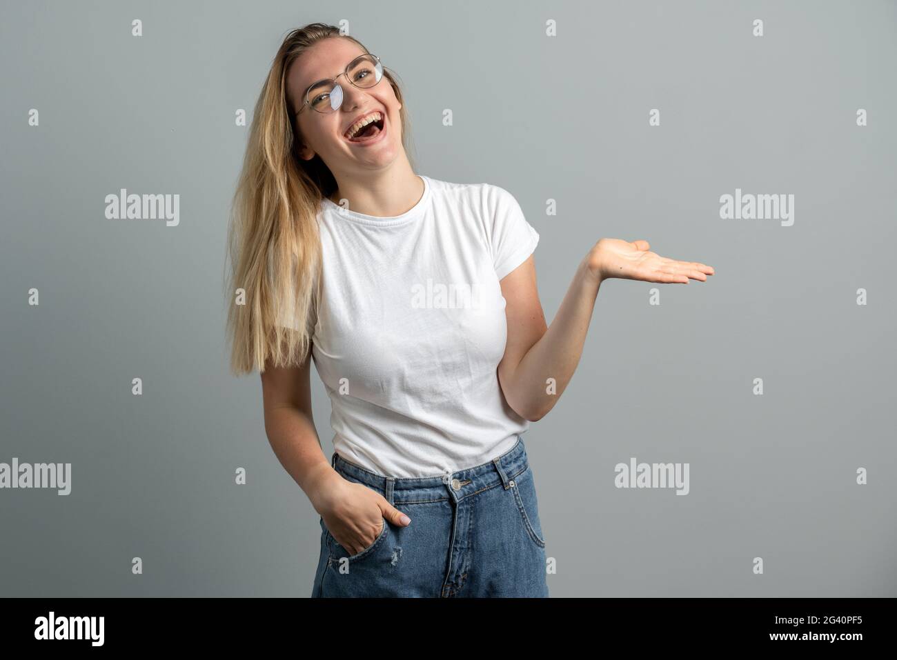 Emotionale Blondine mit Brille, die auf grauem Hintergrund laut lächelt. Schönes Mädchen mit langen Haaren auf einem Hintergrund der leeren Wand. Stockfoto