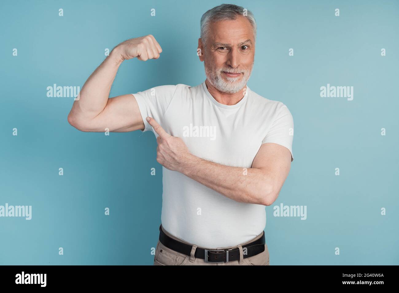 Älterer bärtiger Mann, der Muskeln in weißer T-Shirt-Pose auf blauem Studiohintergrund zeigt. Ernsthafter Mann weist auf seinen perfekten Bizeps hin Stockfoto