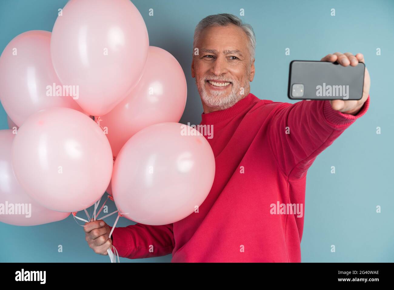 Fröhlicher, lächelnder, grauhaariger Mann mit Kugeln auf blauem Hintergrund. Positive Person, die ein Selfie am Telefon macht, fröhlich, lächelnd, grauhaariger Mann w Stockfoto