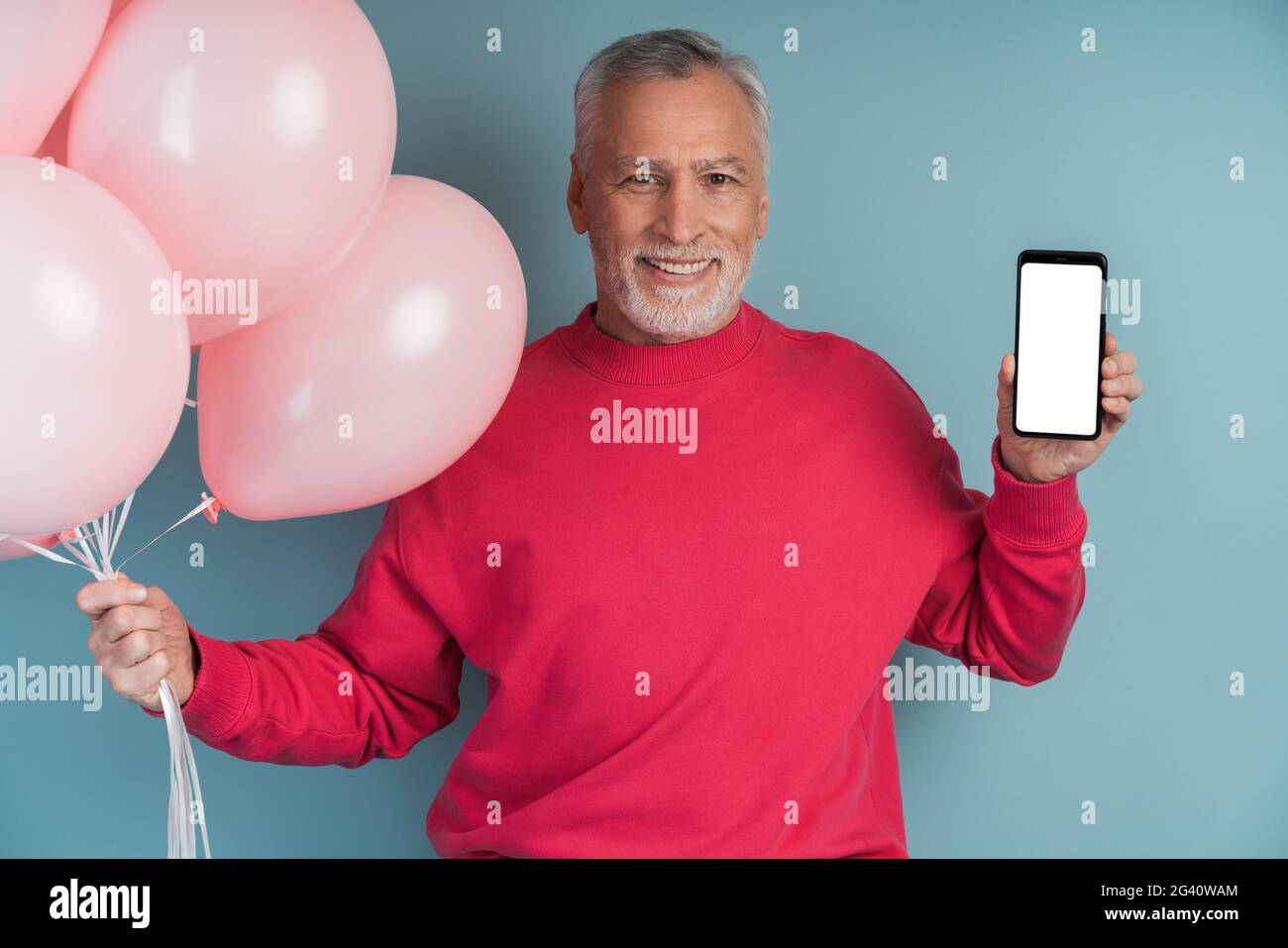 Mann mit Luftballons, die einen hellen Pullover tragen und auf blauem Hintergrund posieren. Netter Mann, der ein Telefon hält, einen leeren, schwarzen Telefonbildschirm zeigt, Platz zum Kopieren, Platz Stockfoto