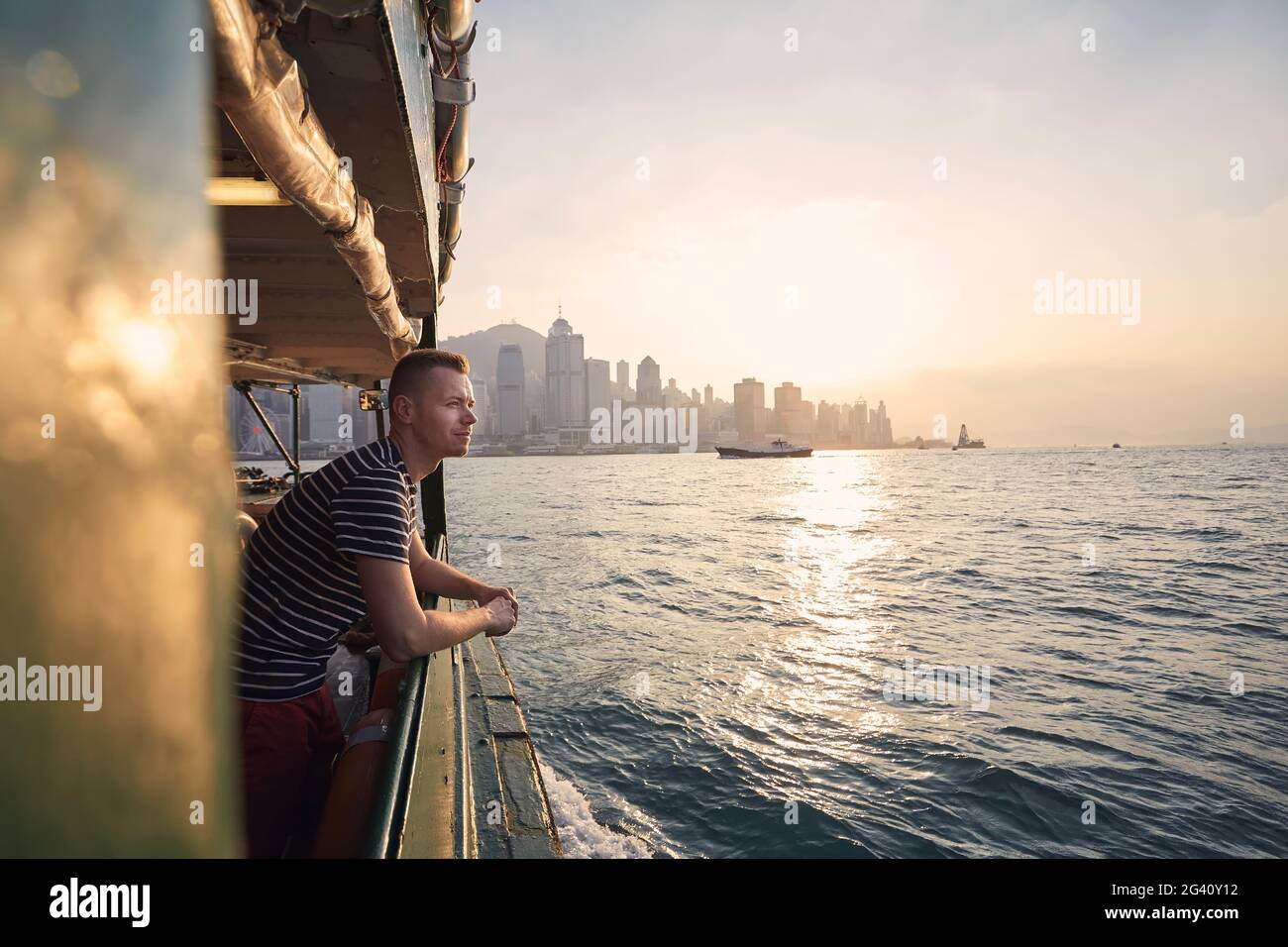 Nachdenklicher Mann, der vom Fährschiff aus auf die Skyline der Stadt blickt und einen wunderschönen Sonnenuntergang genießt. Touristen in Hongkong. Stockfoto