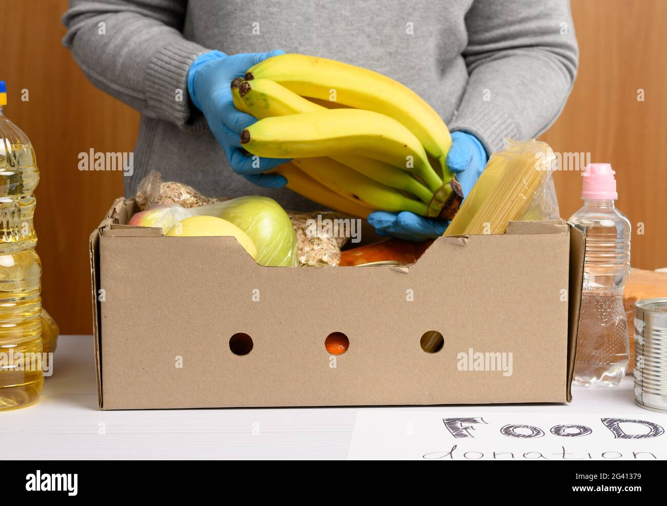 Frau in Handschuhen sammelt immer wieder Lebensmittel, Früchte und Dinge und einen Karton, um Bedürftigen zu helfen Stockfoto