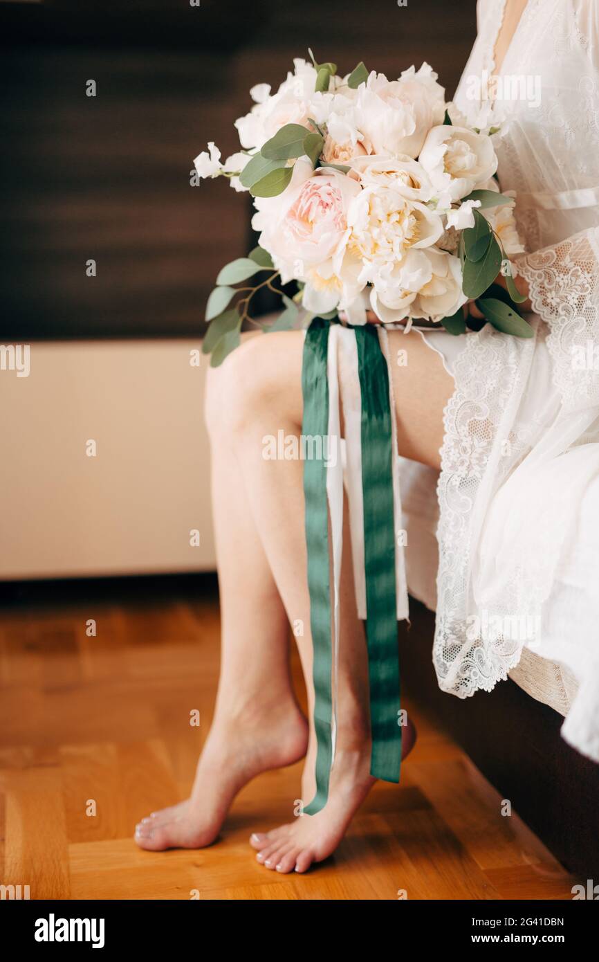 Brautstrauß aus cremefarbenen Rosen und Eukalyptus, der während der Vorzeremonie auf den Beinen der Braut liegt Stockfoto