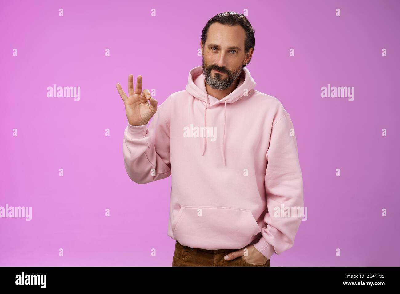 Selbstbewusst gut aussehende glücklich alten Macho Kerl bärtig graue Haare in stilvollen rosa Kapuzenpullover halten Handtasche zeigen okay ok Genehmigung gestu Stockfoto