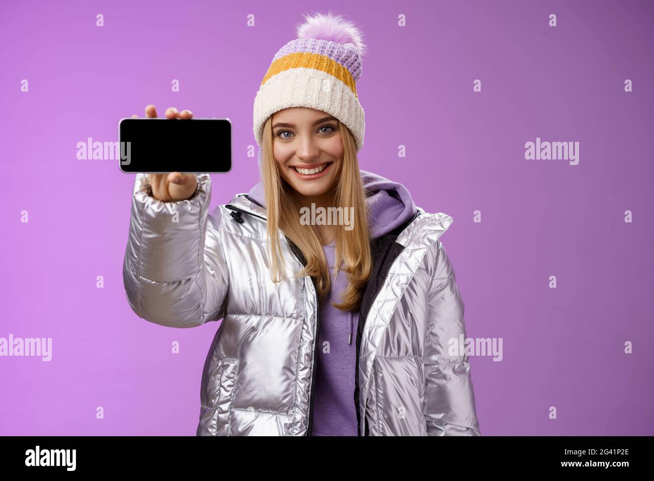 Zuversichtlich freundlich gut aussehende blonde Mädchen im Freien Silber glitzernden Jacke Hut halten Smartphone horizontal zeigt mobile pho Stockfoto