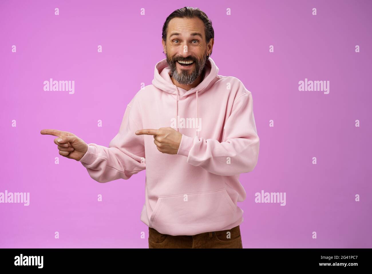 Charismatischer, fröhlicher, gut aussehender, reifer 50er Jahre-Mann mit stylischem Hipster-Hoodie, der aufgeregt lächelt und nach links zeigt, beeindruckt mit seinen Zeigefingern Stockfoto
