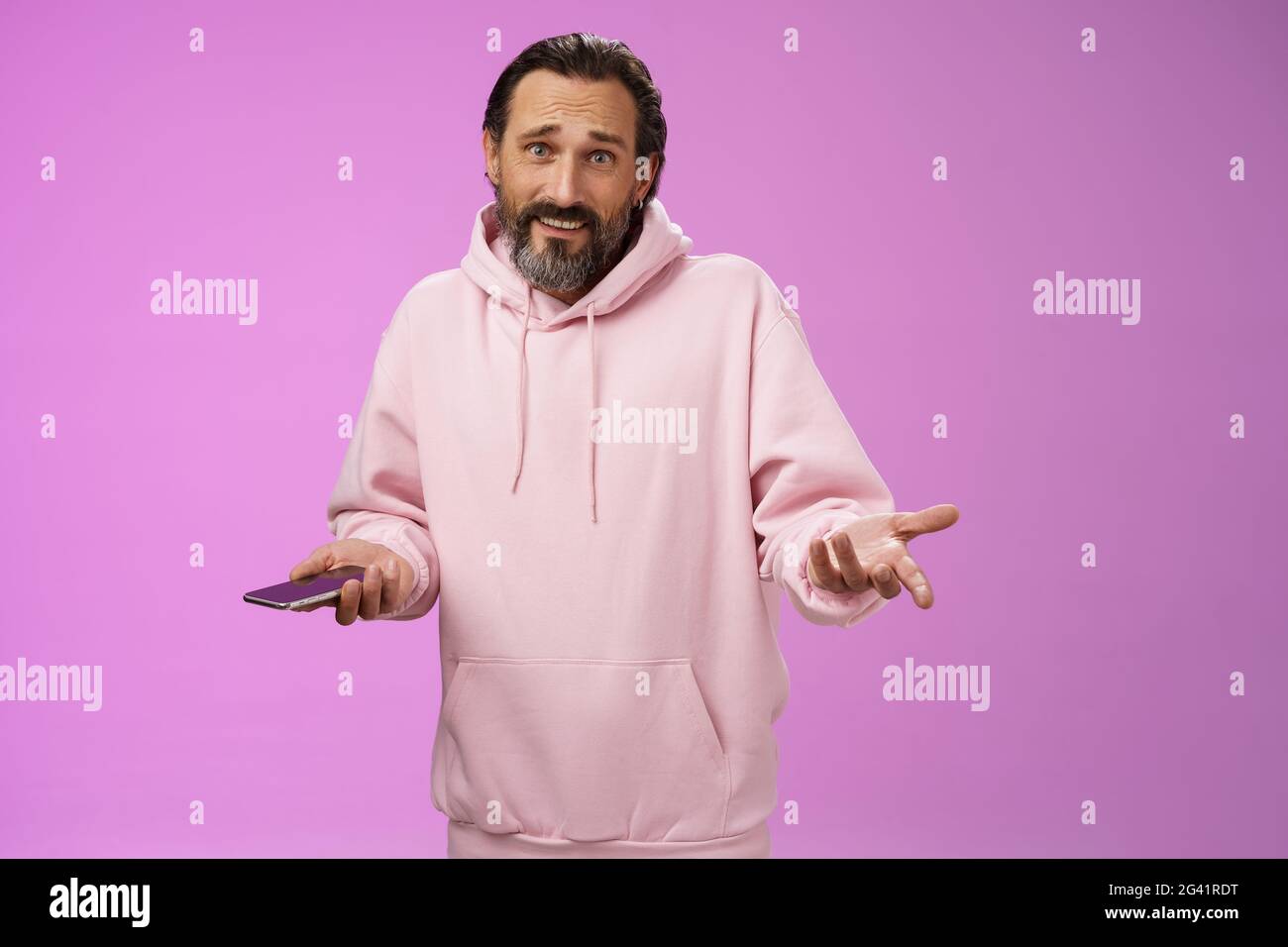 Was soll es denn? Verwirrt befragten Erwachsenen bärtigen stilvollen Kerl in rosa Kapuzenpullover zuckende Hände verbreiten seitwärts Bestürzung halten Smartphone Stockfoto