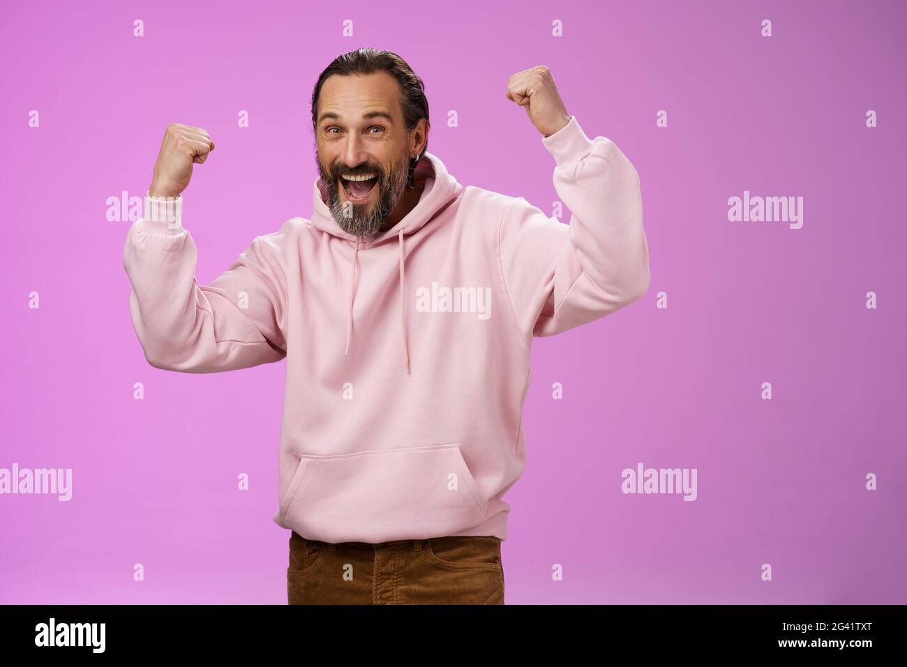 Ja, wer kümmert sich um das Alter. Sorglos glücklich glücklich alten Mann bärtig in trendigen rosa Kapuzenpullover heben Fäuste freudig triumphieren mit Spaß gl Stockfoto
