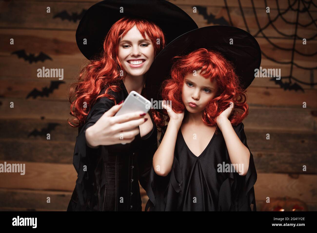 Halloween-Konzept - schöne kaukasische Mutter und ihre Tochter mit langen roten Haaren in Hexen-Kostümen, die ein Selfie mit smartp machen Stockfoto