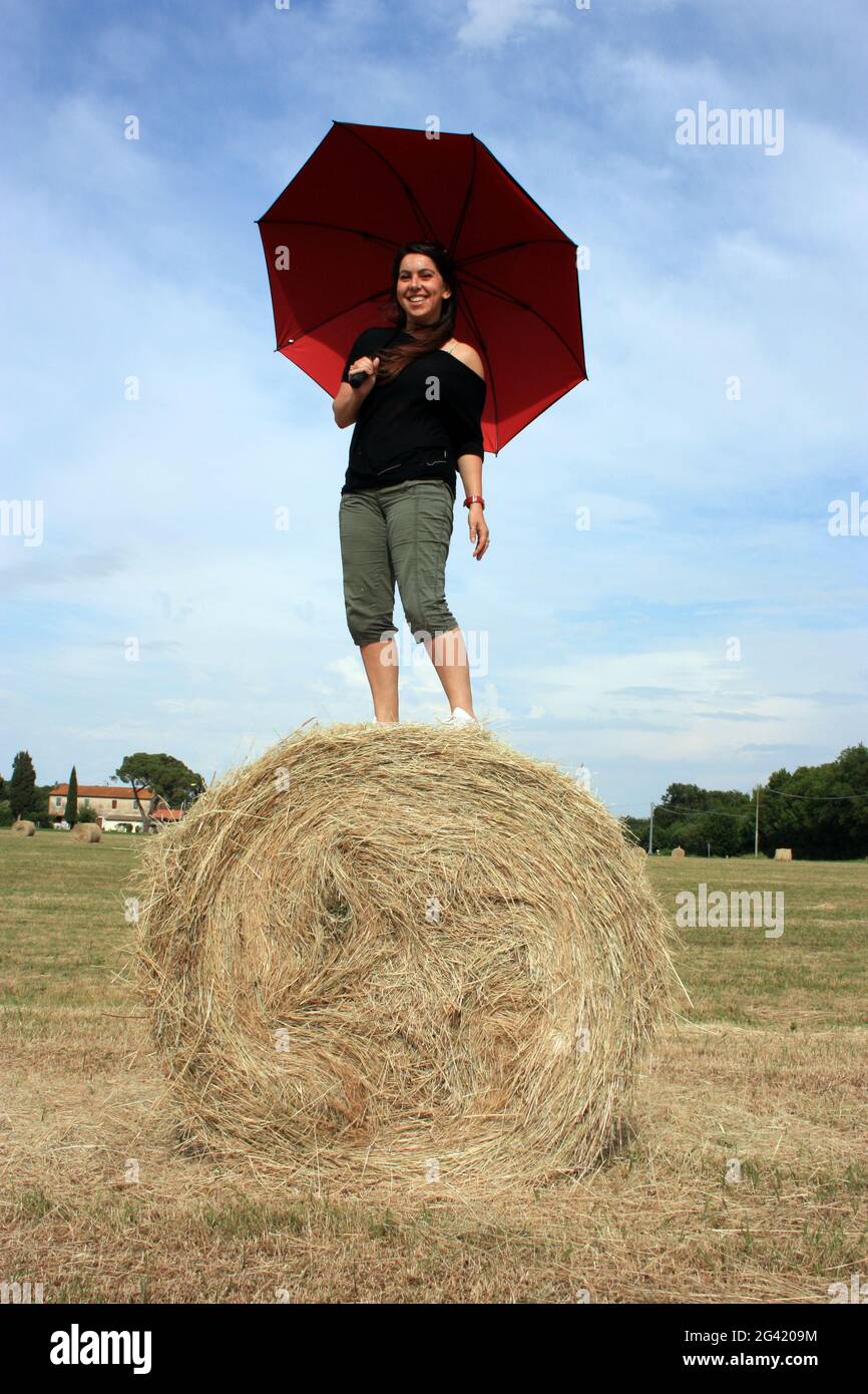 Junge Frau auf einem Heuball in einem gepflügten Feld in der toskana im Sommer unter dem Sonnenschirm genießen Stockfoto