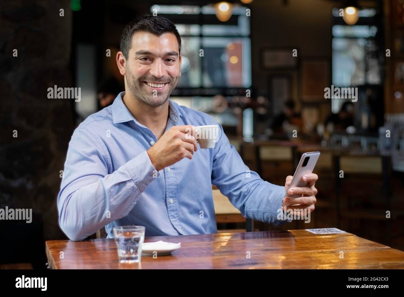 Junger latein in einem Café, sitzt an einem Tisch, trinkt Kaffee und überprüft sein Smartphone Stockfoto