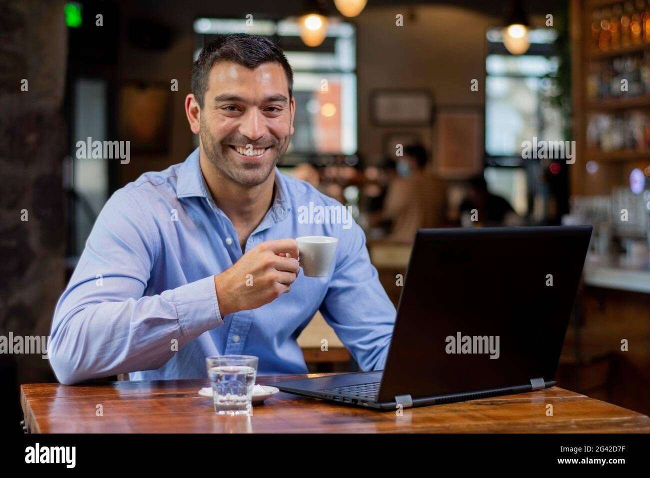 Ein junger latein in einem Café, der an einem Tisch sitzt, Kaffee trinkt und sein Smartphone überprüft, lächelnd auf die Kamera schaut Stockfoto