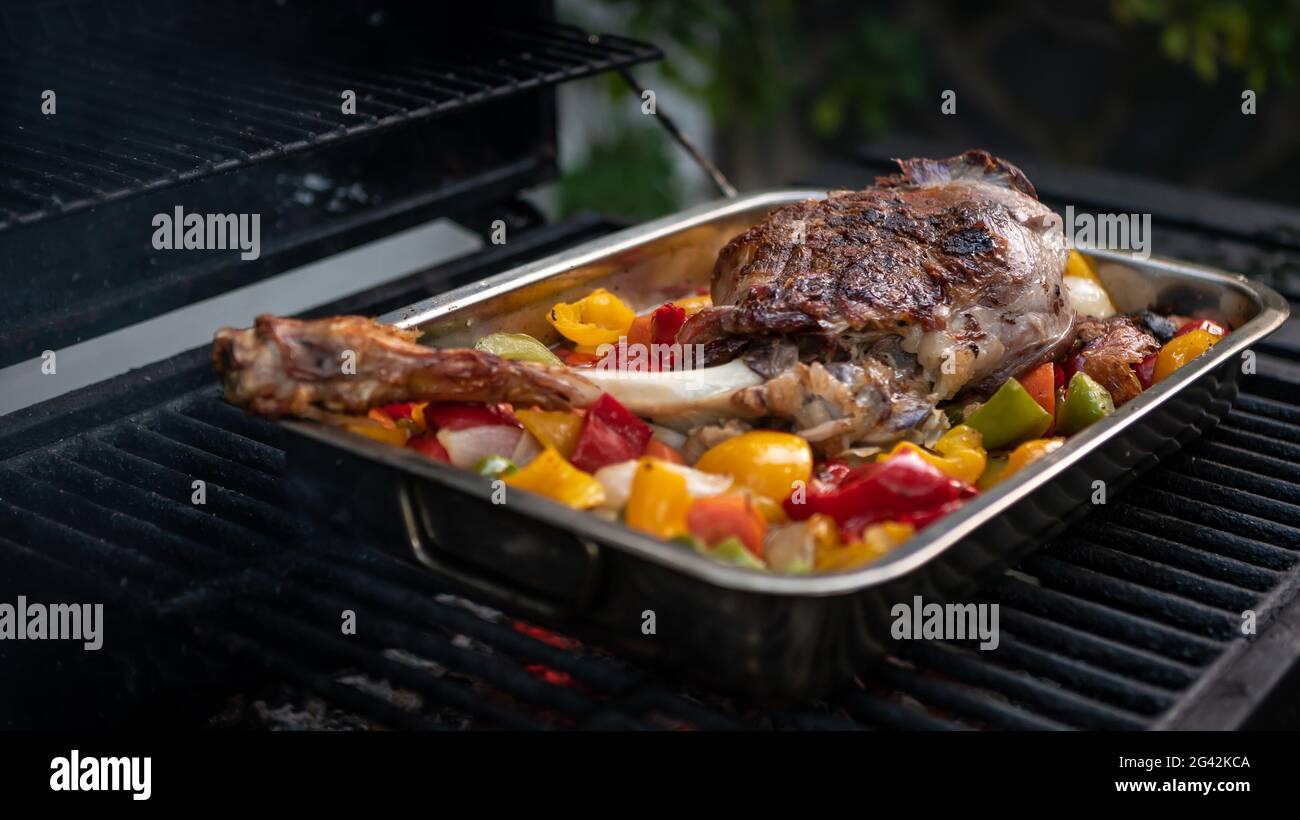 Koch kocht ein köstliches Lammkeule auf einem Grill mit farbigen Paprika und Kartoffeln in einem Metalltablett. Nahaufnahme von leckerem Ziegenfleisch auf dem Grill des Gartenhauses Stockfoto