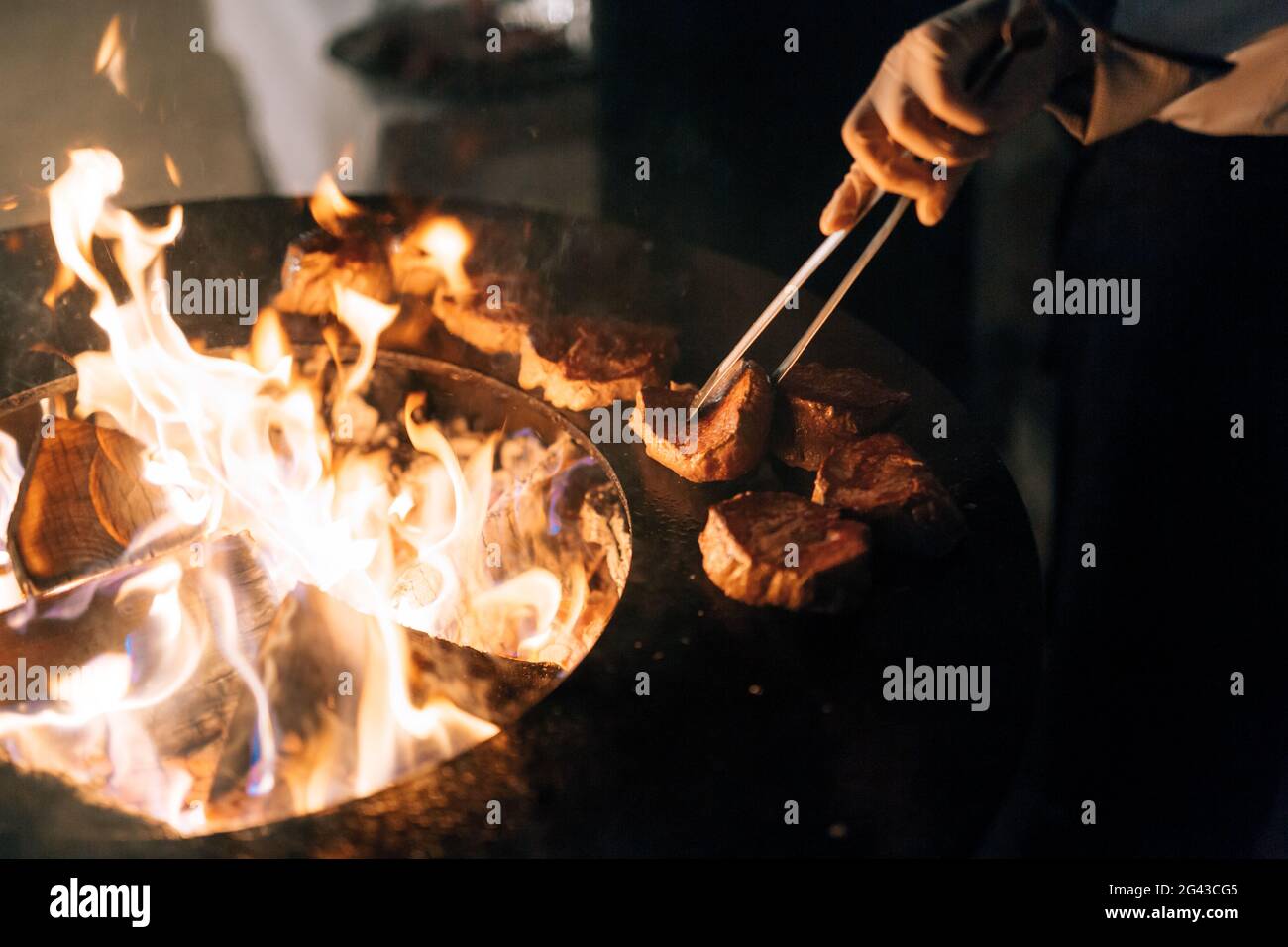 Der Koch grilbert Steaks. Das Fleisch mit einer Zange wenden. Gegrillter runder Tisch mit einem Feuer im Inneren. Stockfoto