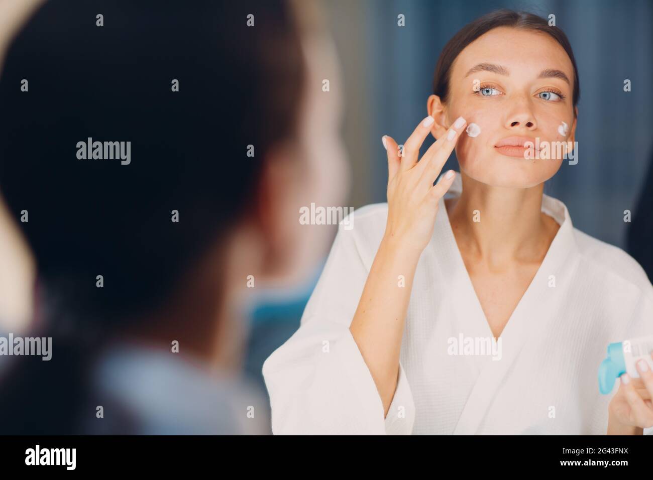 Junge Frau, die am Spiegel kosmetische weiße Creme auf ihr Gesicht appied Stockfoto
