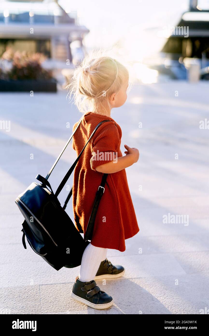 Ein süßes 2-jähriges Kind trägt einen großen schwarzen Rucksack Auf ihrem Rücken, während sie auf einem Bootssteg spazieren Stockfoto