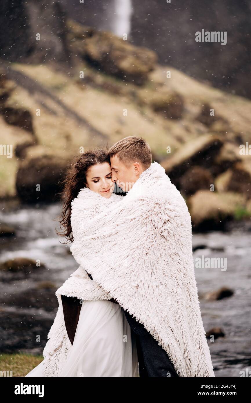 Ein Hochzeitspaar steht am Ufer eines Bergflusses unter einer Wolldecke. Der Bräutigam umarmt die Braut und versucht sie zu wärmen. S Stockfoto