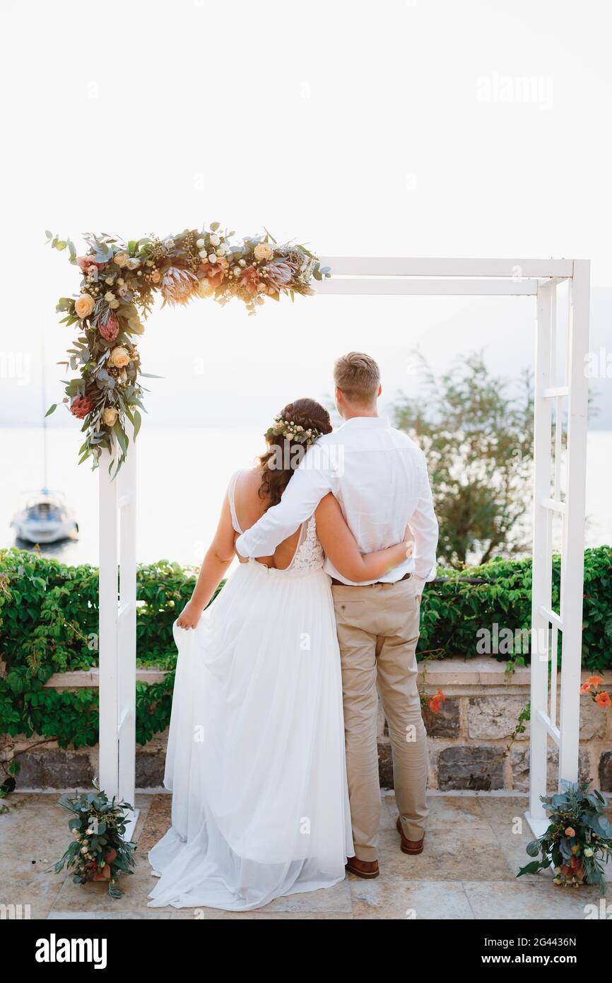Die Braut und der Bräutigam stehen umarmt unter dem Hochzeitstor und blicken auf das Meer, Rückansicht Stockfoto