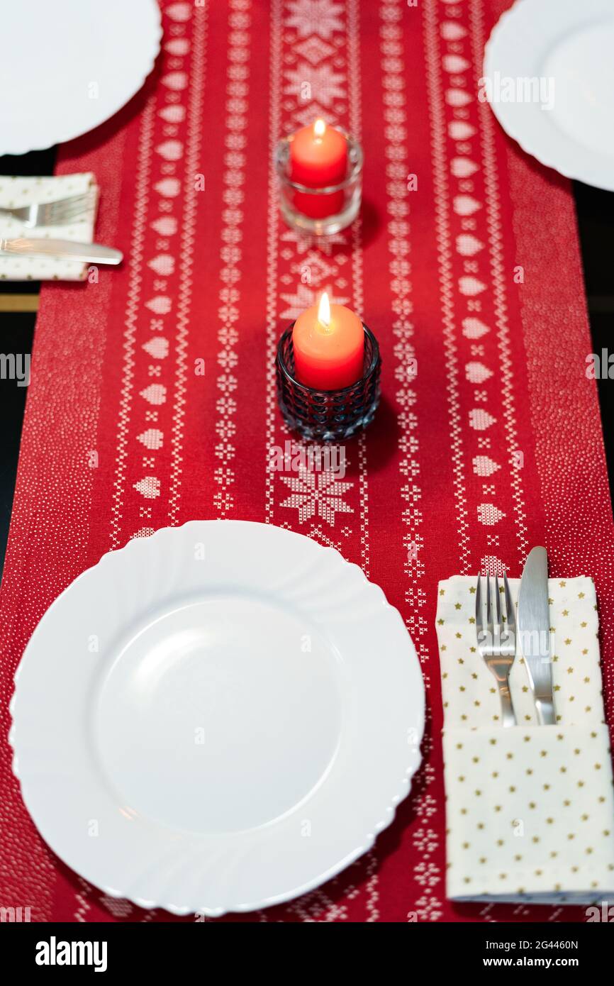 Festliche Tischdekoration für die Neujahrsfeiertage mit roten Kerzen und weißen Tellern auf einer roten Stofftischdecke mit patte Stockfoto