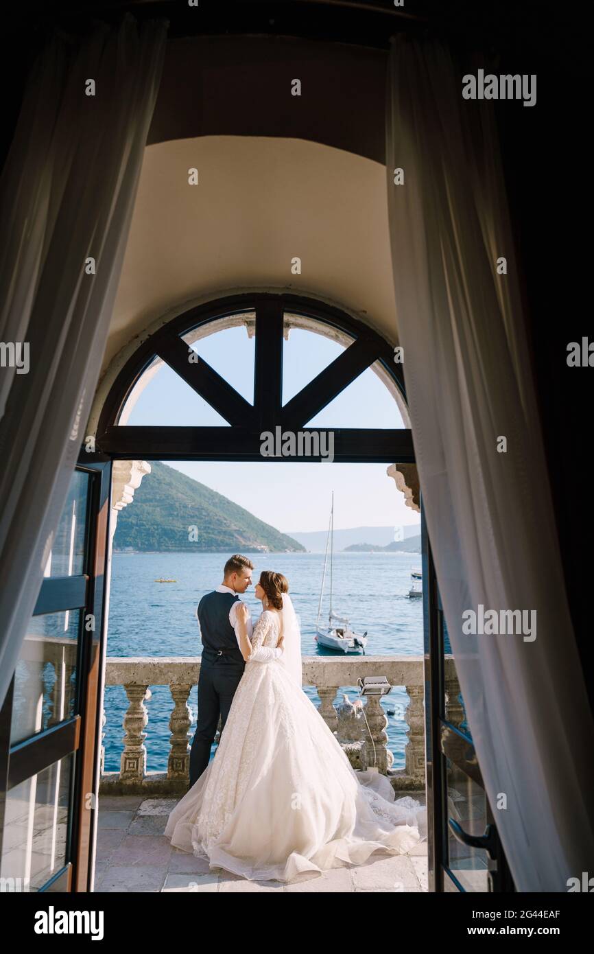 Das Hochzeitspaar steht auf einem Hotelbalkon mit Meerblick, Blick durch ein offenes antikes Fenster. Hochzeitsfoto der feinen Kunst in Montenegro Stockfoto