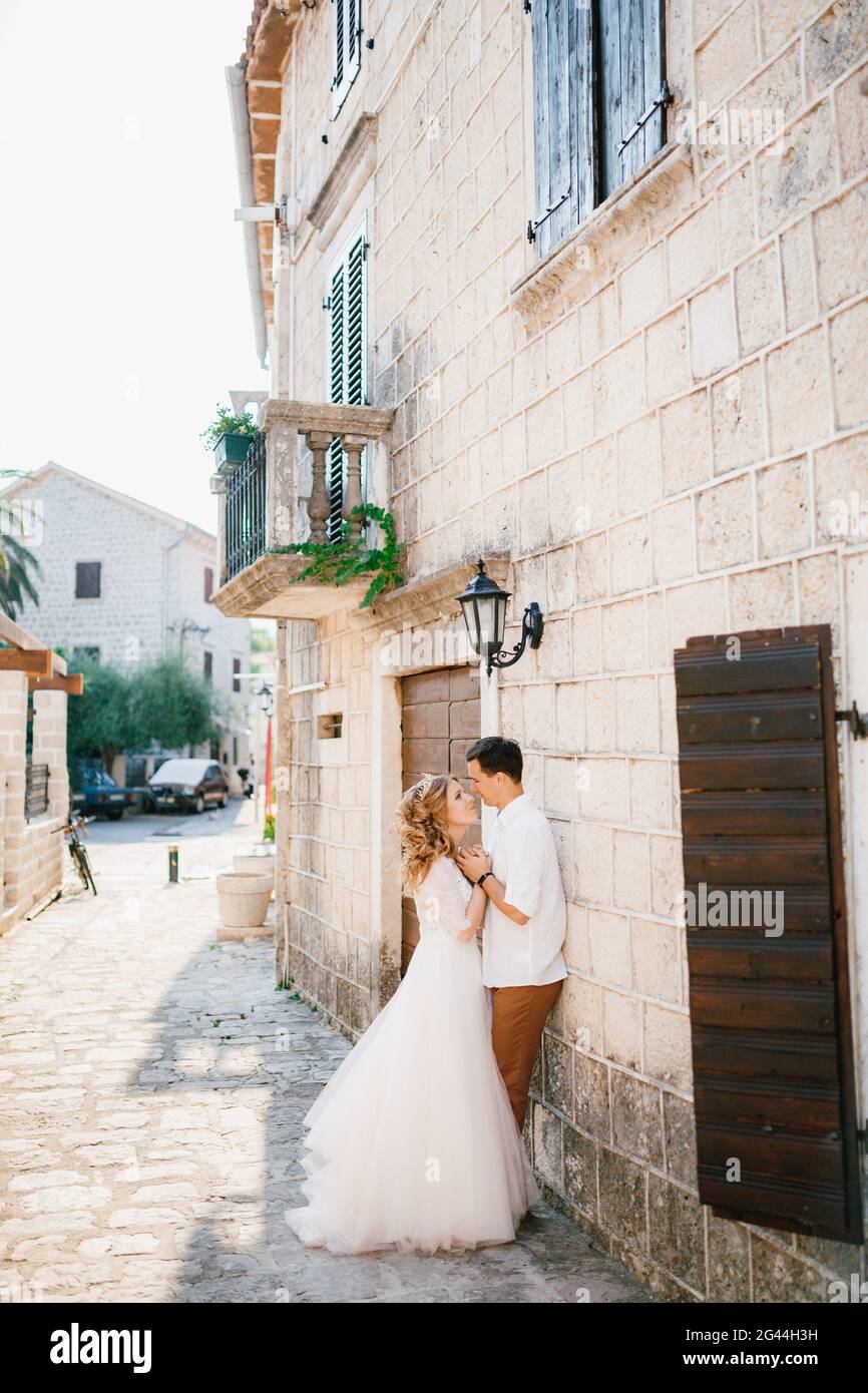 Braut und Bräutigam umarmen sich in der Nähe eines schönen weißen Gebäudes mit Balkonen und hölzernen Fensterläden in der Altstadt von Perast in der Nähe eines jungen TR Stockfoto
