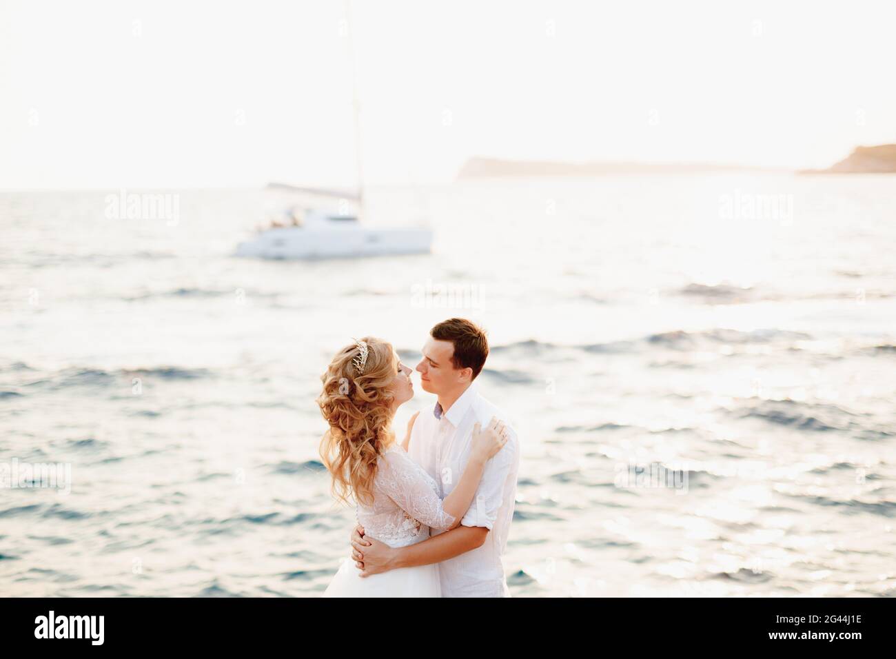 Die Braut und der Bräutigam umarmen sich am Meer und sind dabei, sich zu küssen, hinter ihnen segelt eine weiße Yacht Stockfoto