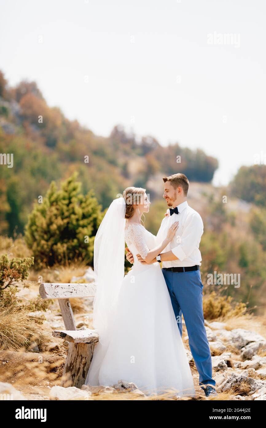 Braut und Bräutigam umarmen sich zärtlich in der Nähe eines Holzes Bank in den Bergen und schauen sich gegenseitig an Stockfoto
