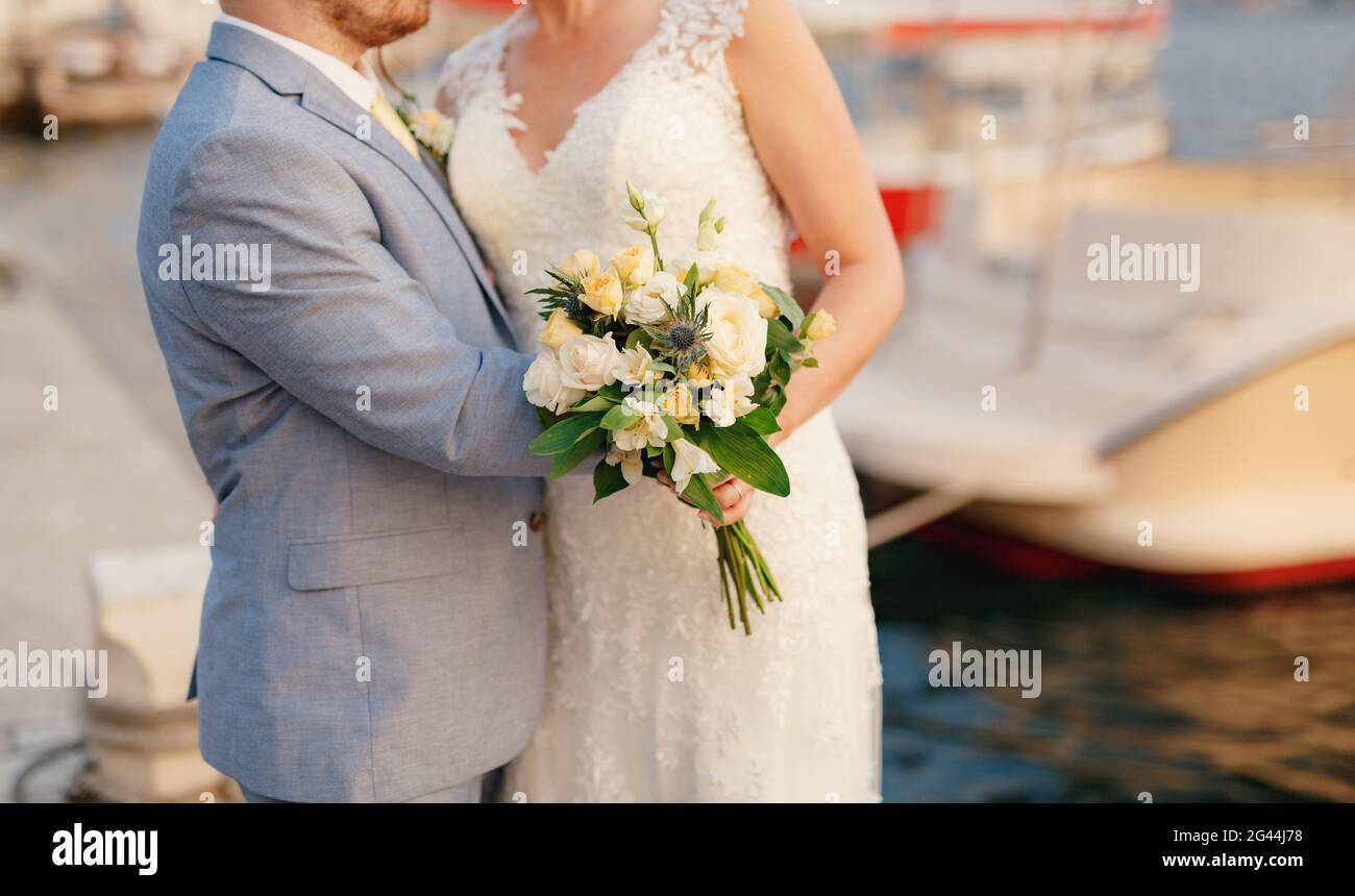 Braut und Bräutigam umarmen sich am Pier am Wasser, hinter ihnen sind Boote, die Braut hält einen Strauß Stockfoto