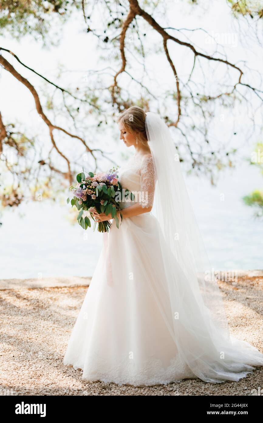 Eine raffinierte Braut steht mit einem Brautstrauß in ihr Hände unter den Ästen eines Baumes am Meer Stockfoto
