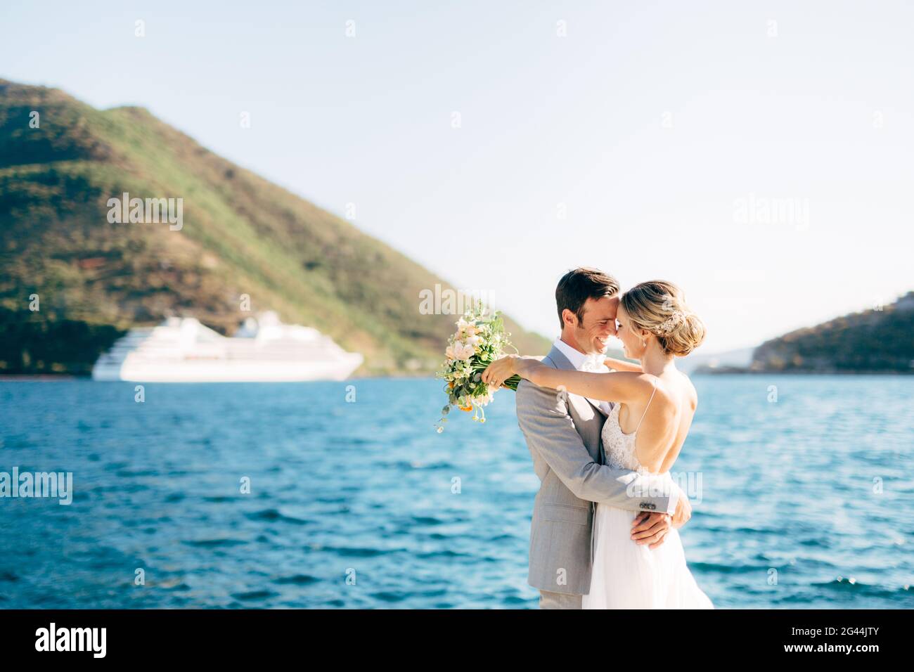 Braut und Bräutigam umarmen sich am Pier in der Bucht von Kotor, hinter ihnen sind grüne Berge und ein weißes Touristenschiff Stockfoto