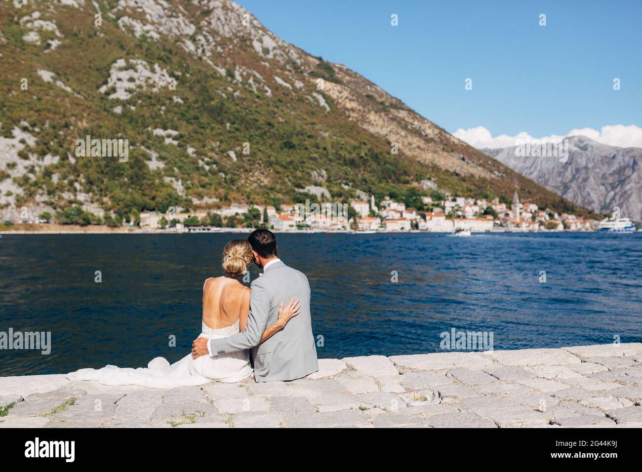 Braut und Bräutigam sitzen umarmt am Pier in der Bucht von Kotor, vor ihnen ist die Altstadt Stockfoto
