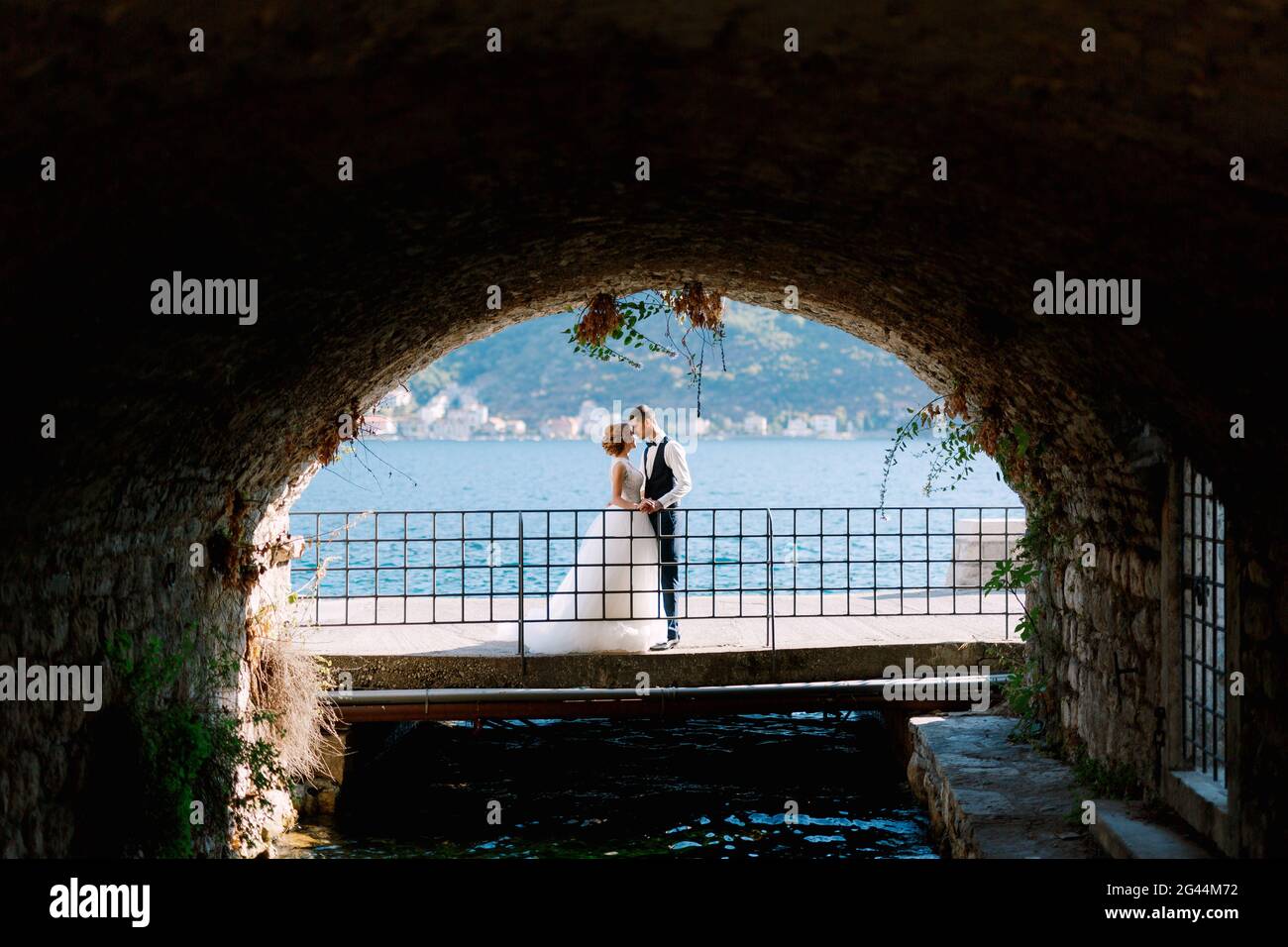 Die Braut und der Bräutigam umarmen und küssen sich auf der Brücke unter dem Bogen, hinter ihnen ist die Bucht von Kotor Stockfoto